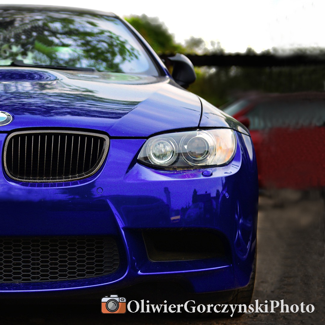 Fragment niebieskiego BMW M3 E92 z widoczną kratką grilla, reflektorami i fragmentem maski, z rozmyciem tła i logo fotografa w dolnej części kadru.