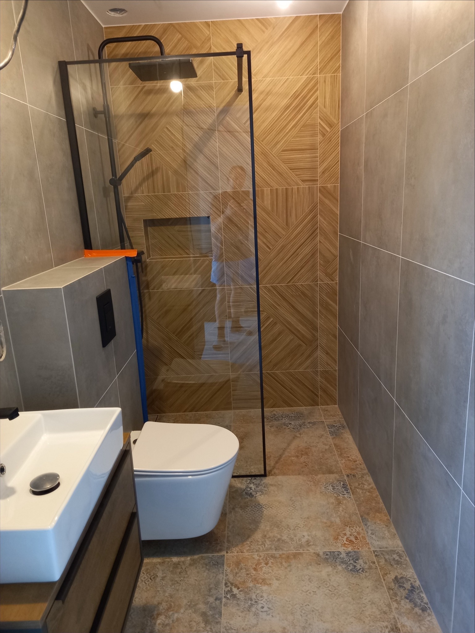 Wykończona łazienka z kabiną prysznicową typu walk-in, umywalką nablatową na szafce z drewnianym frontem i podwieszaną toaletą; na ścianach i podłodze różne rodzaje płytek.