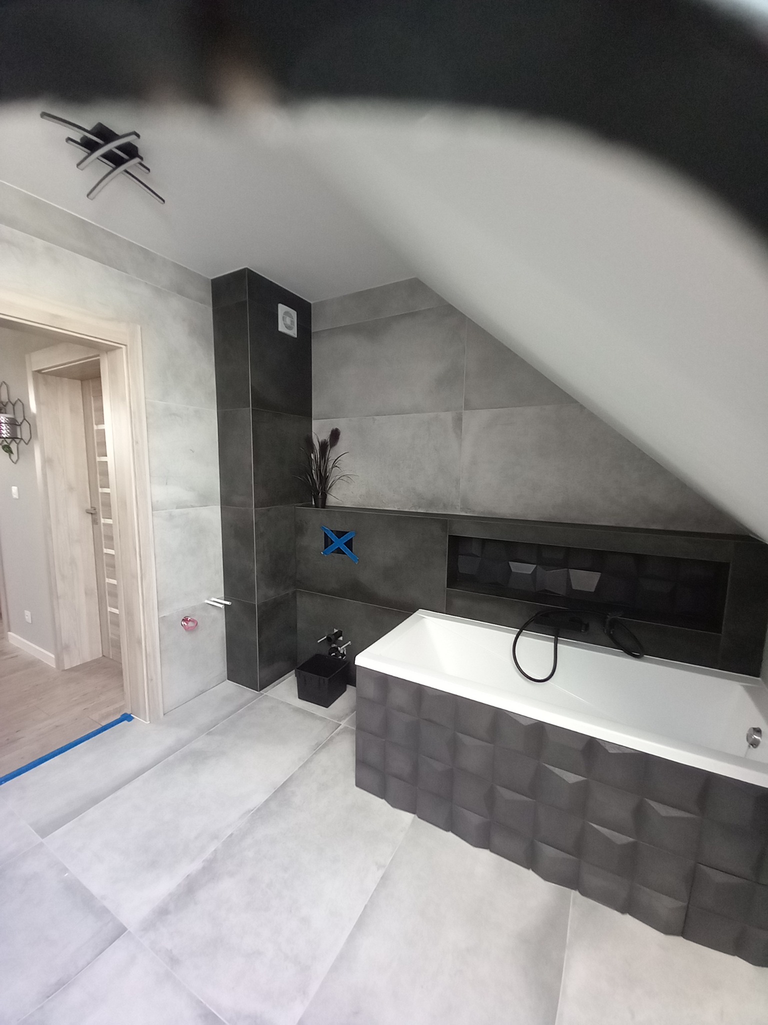 Nowoczesna łazienka na poddaszu z szarymi płytkami imitującymi beton, białą wanną obudowaną ciemnoszarymi panelami 3D, czarną armaturą i dekoracyjną rośliną w tle, widoczne zabezpieczenia na ścianie.