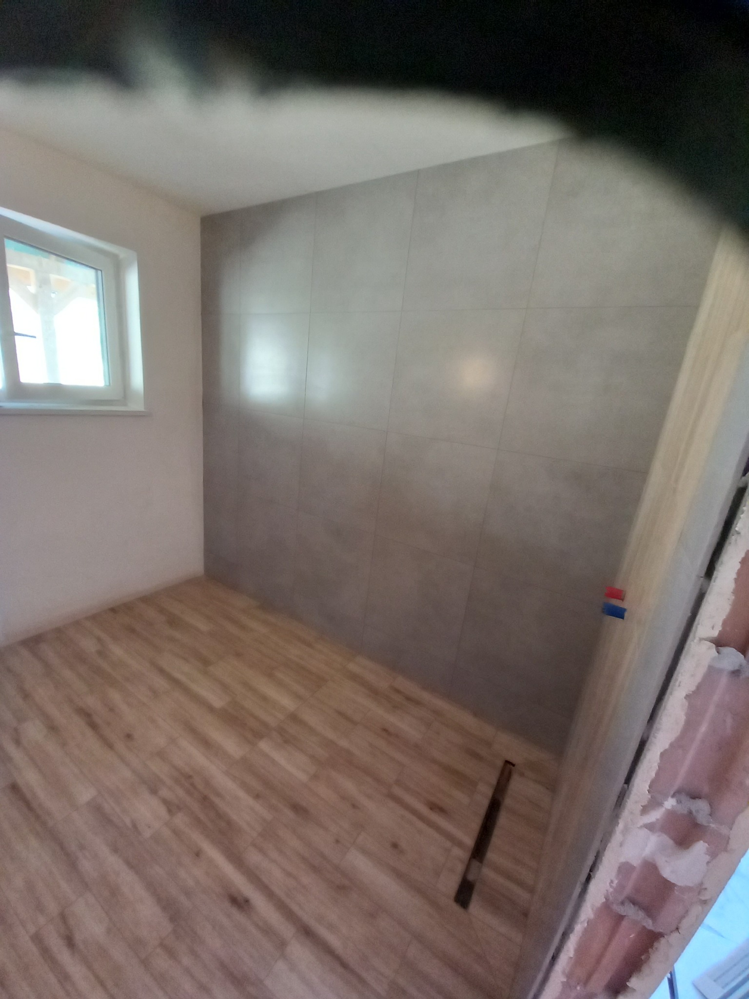 Wnętrze łazienki z ułożonymi szarymi płytkami na ścianie i panelami podłogowymi imitującymi drewno, widoczny odpływ liniowy w podłodze i fragment niedokończonej obudowy drzwi.