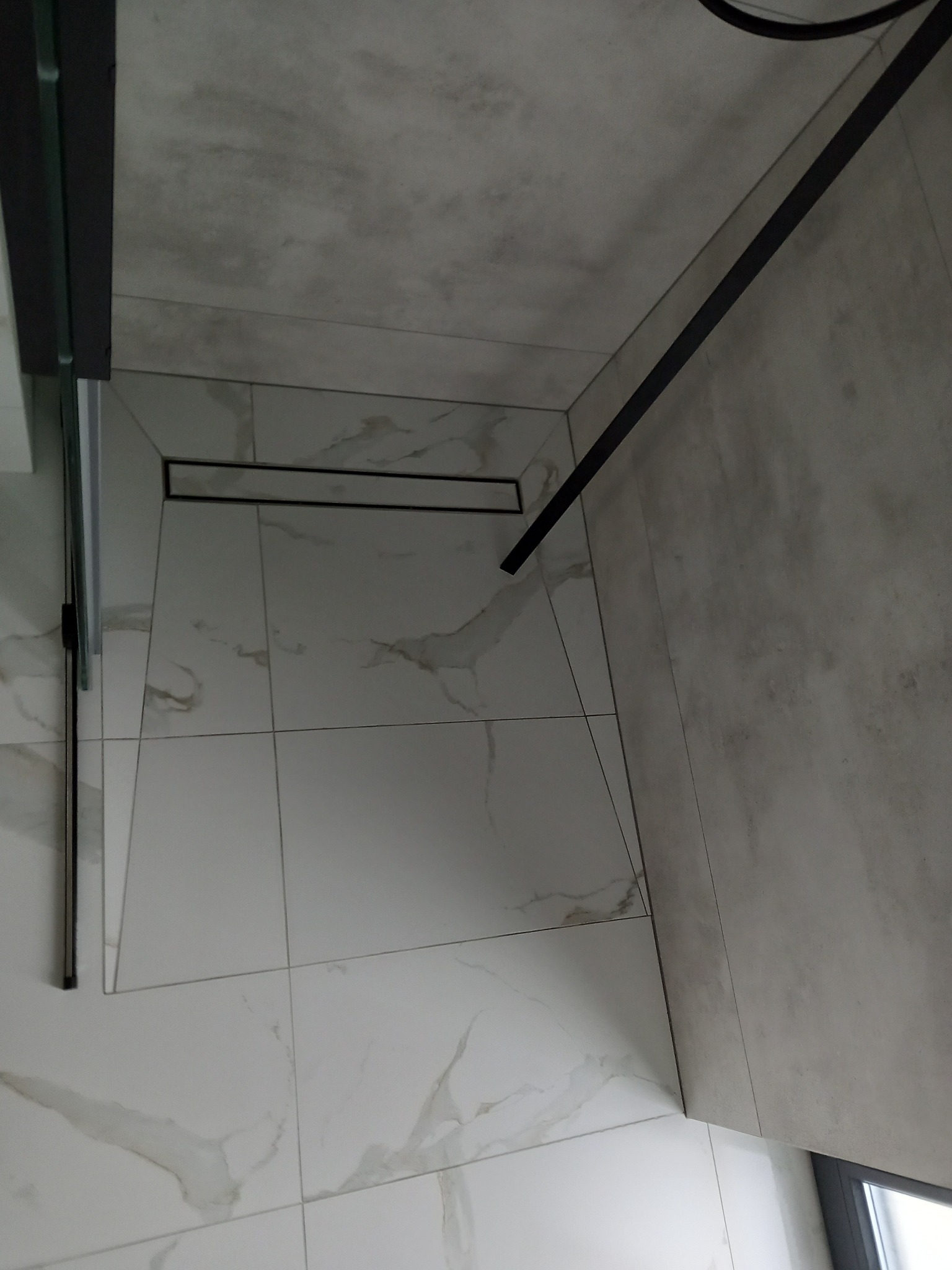 Białe płytki podłogowe z marmurkowym wzorem w kabinie prysznicowej z czarnym odpływem liniowym i szklanymi drzwiami.