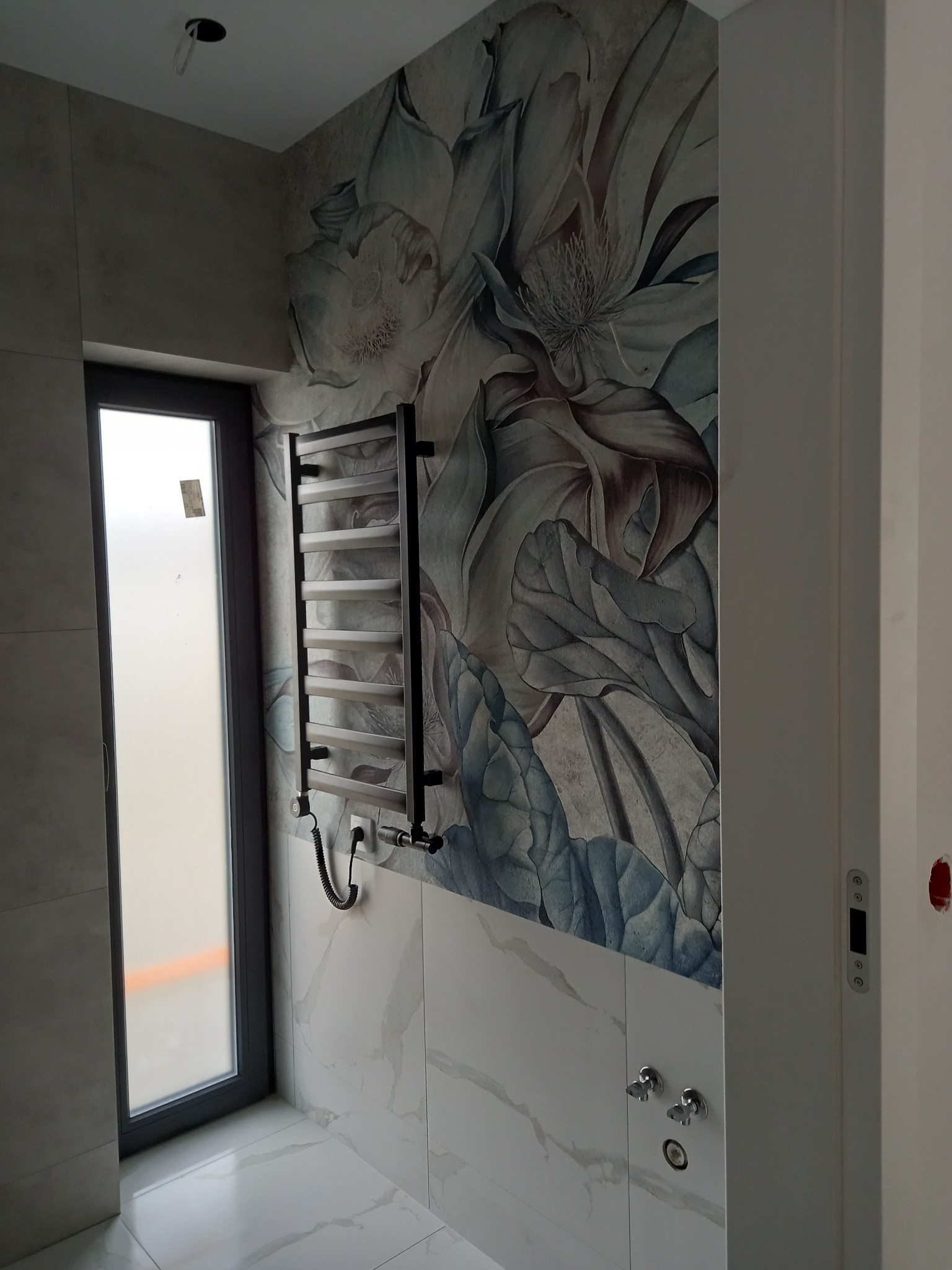Wykończona łazienka z grzejnikiem drabinkowym na ścianie z motywem kwiatowym, marmurowe płytki na ścianie i podłodze, widoczne zawory i okno.