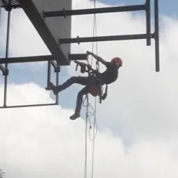 Pracownik w kasku i uprzęży, zawieszony na linach, wykonuje prace na wysokościach na konstrukcji stalowej, z widocznym fragmentem nieba z chmurami w tle.