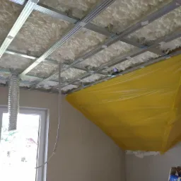 Poddasze w trakcie izolacji pianą, widoczna metalowa konstrukcja pod przyszłą zabudowę, folia ochronna na ścianie, fragment okna w tle.