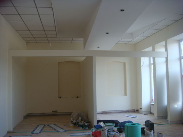 Wnętrze w trakcie remontu: widoczne elementy konstrukcyjne sufitu podwieszanego, świeżo pomalowane ściany w odcieniach kremu i beżu, podłoga częściowo zabezpieczona folią, farby i narzędzia...