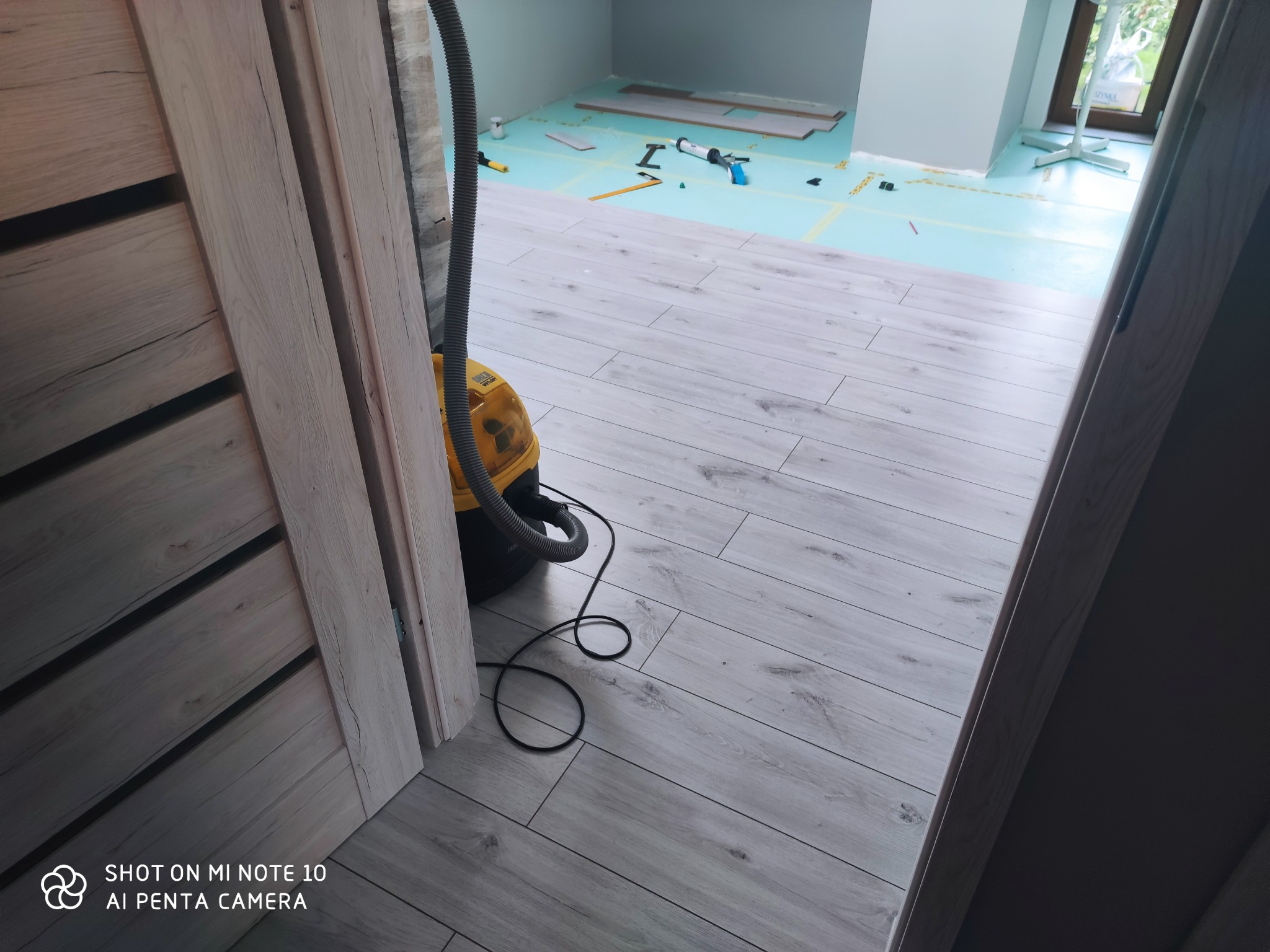 Widok zza uchylonych drzwi na podłogę z paneli w trakcie montażu, widoczny odkurzacz przemysłowy oraz narzędzia na podkładzie pod panele.