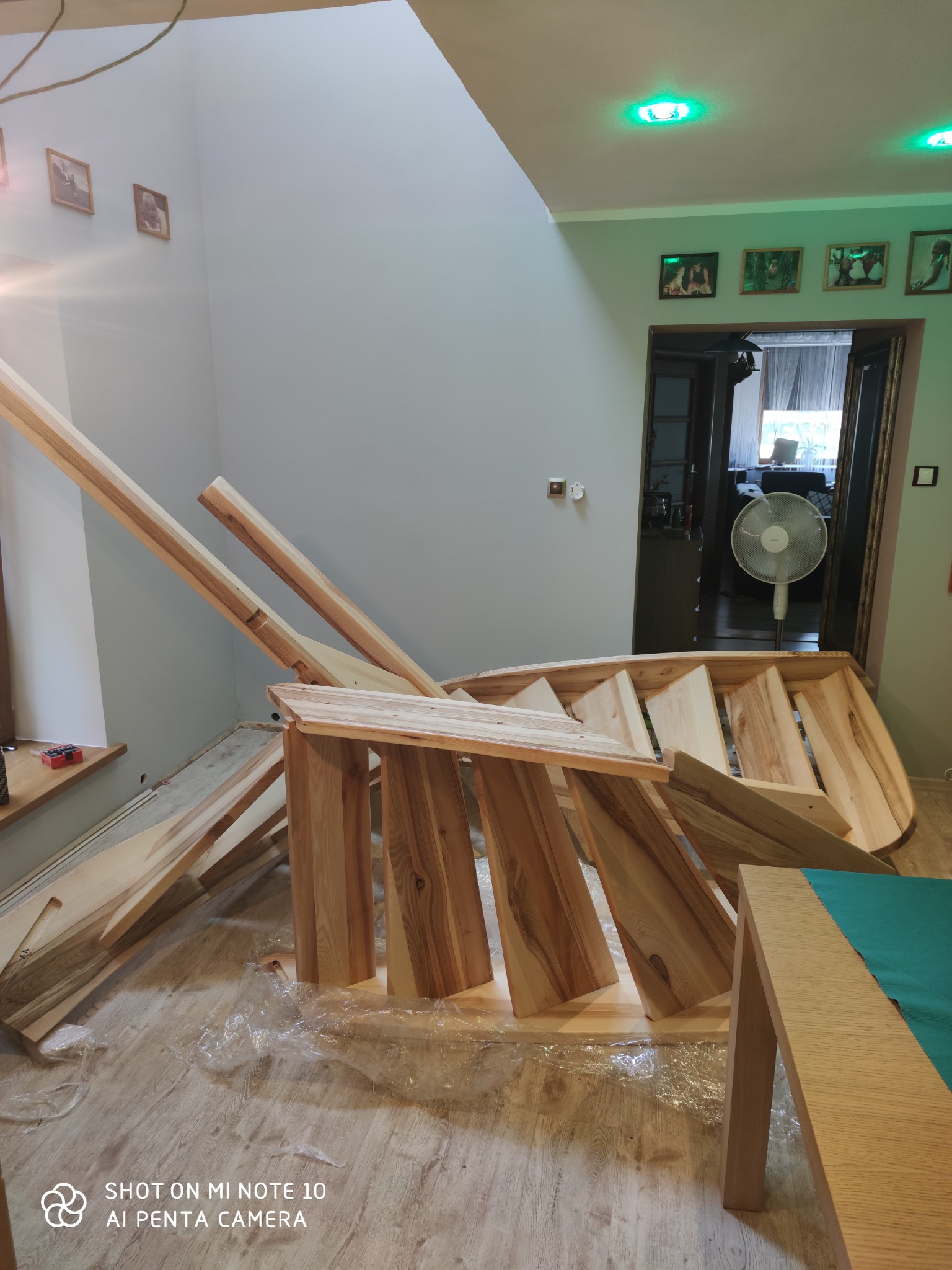 Wnętrze z częściowo zmontowanymi drewnianymi schodami, zabezpieczona podłoga folią, widoczne elementy konstrukcyjne i fragment stołu z zielonym blatem.