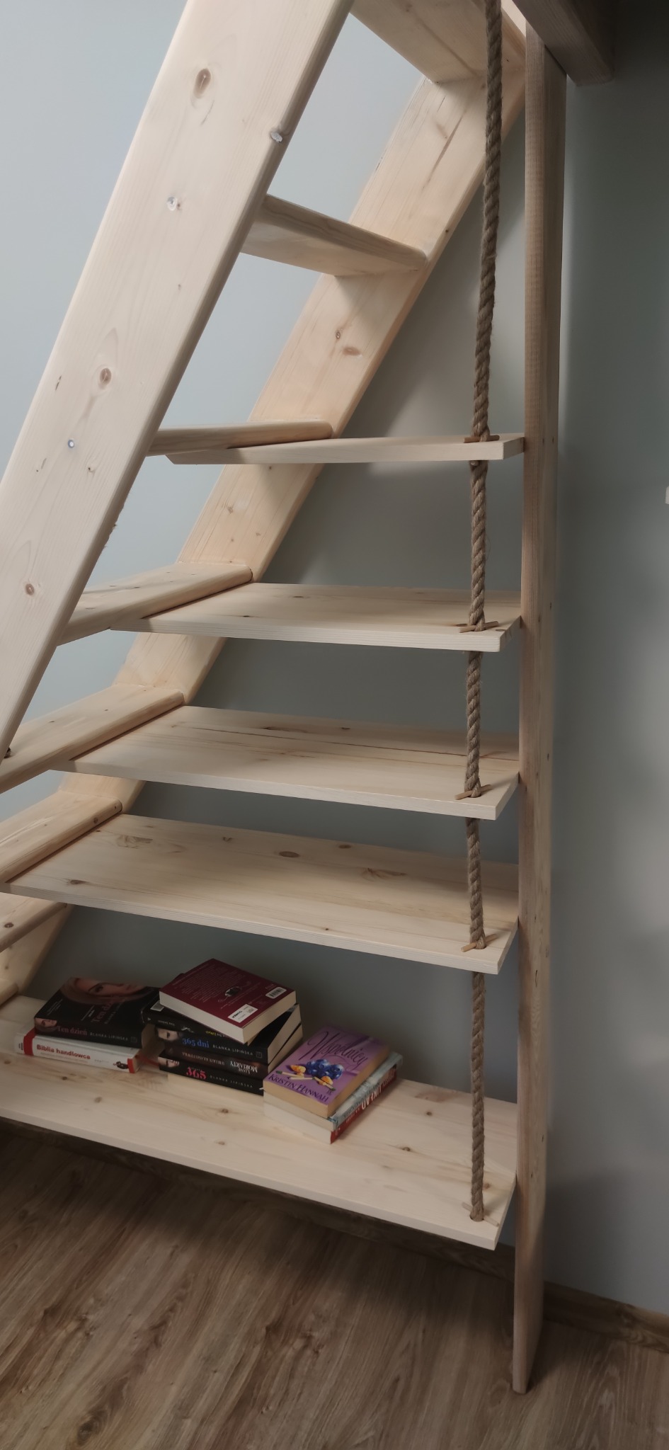 Drewniane schody z półkami na książki, wsparte liną, z widocznymi detalami naturalnego drewna i podłogą w tle.