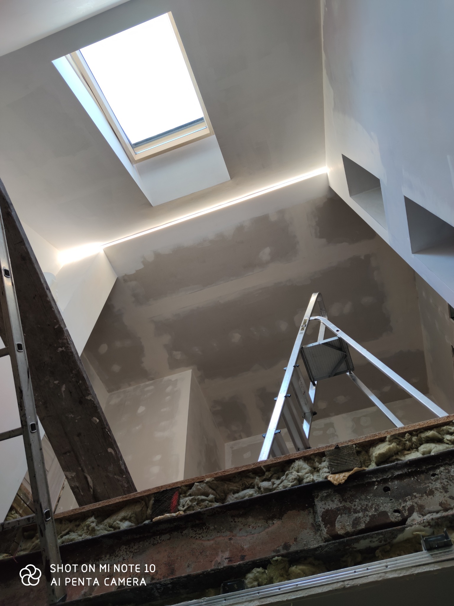 Poddasze w trakcie prac wykończeniowych: widoczna izolacja, metalowa konstrukcja, dwie drabiny, okno dachowe i ściany przygotowane pod malowanie z oświetleniem LED.