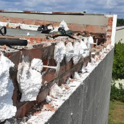 Ocieplanie muru nad dachem z użyciem styropianu mocowanego na kołki, widoczne narzędzia i fragment dachu z papą.