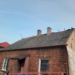 Dom z częściowo zdjętym starym dachem z dachówki ceramicznej, widoczne kominy i fragmenty nowej membrany dachowej, ceglana elewacja budynku.