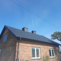 Dom z nowym, grafitowym dachem z blachodachówki, widoczne dwa kominy, rynny i elewacja z cegły, błękitne niebo w tle.