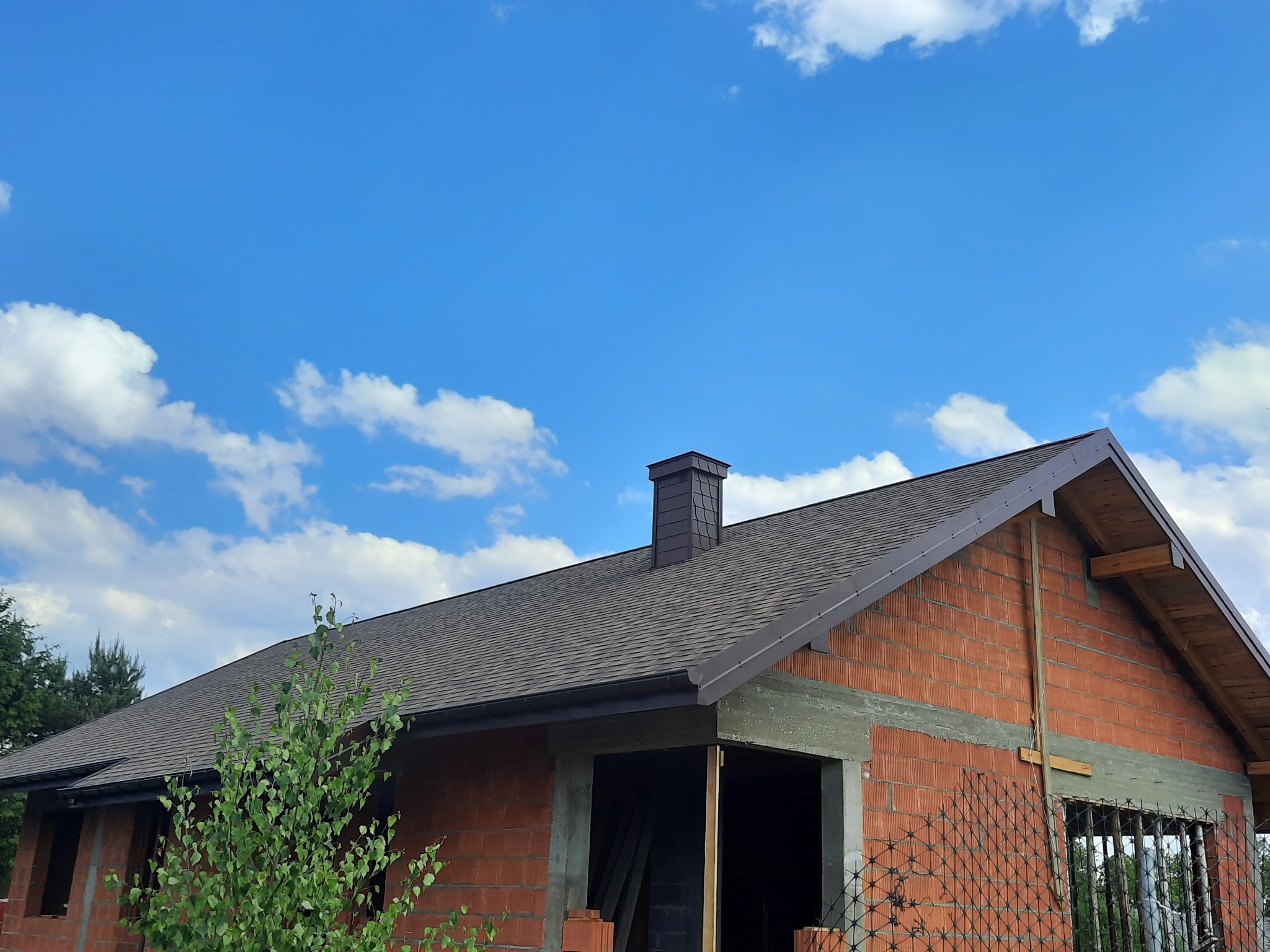 Niedokończony dom z cegły z dachem pokrytym gontem bitumicznym w kolorze grafitowym i kominem z tego samego materiału, na tle błękitnego nieba z białymi obłokami.