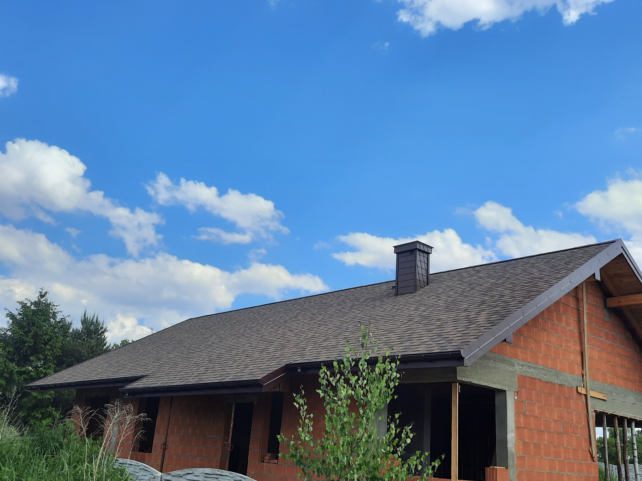 Dom w trakcie budowy z ułożonym brązowym dachem bitumicznym i kominem z blachy w kolorze grafitowym na tle błękitnego nieba z białymi obłokami.