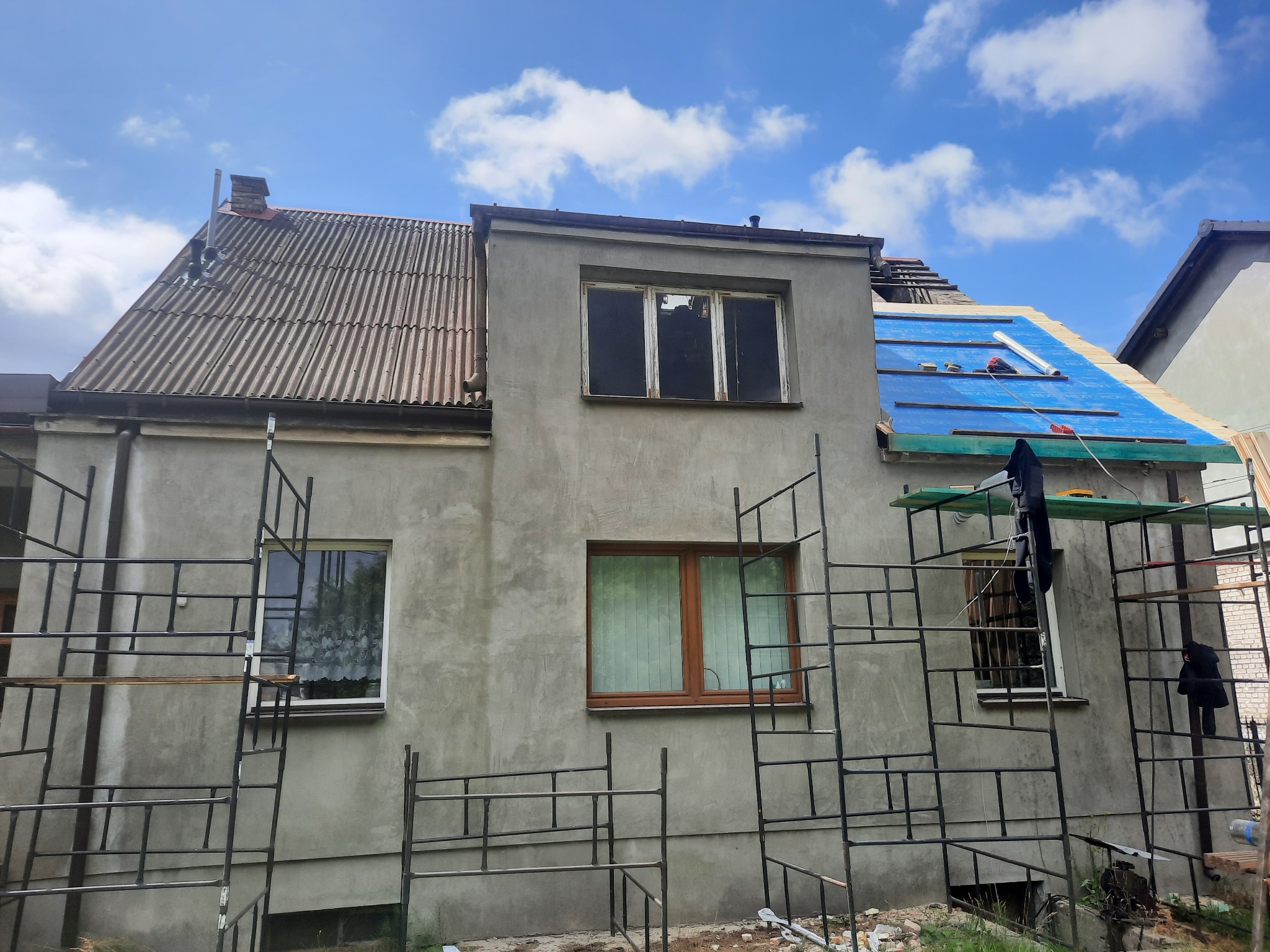 Dom w trakcie wymiany dachu: część dachu pokryta starą dachówką, część zabezpieczona niebieską folią, rusztowanie wokół budynku.