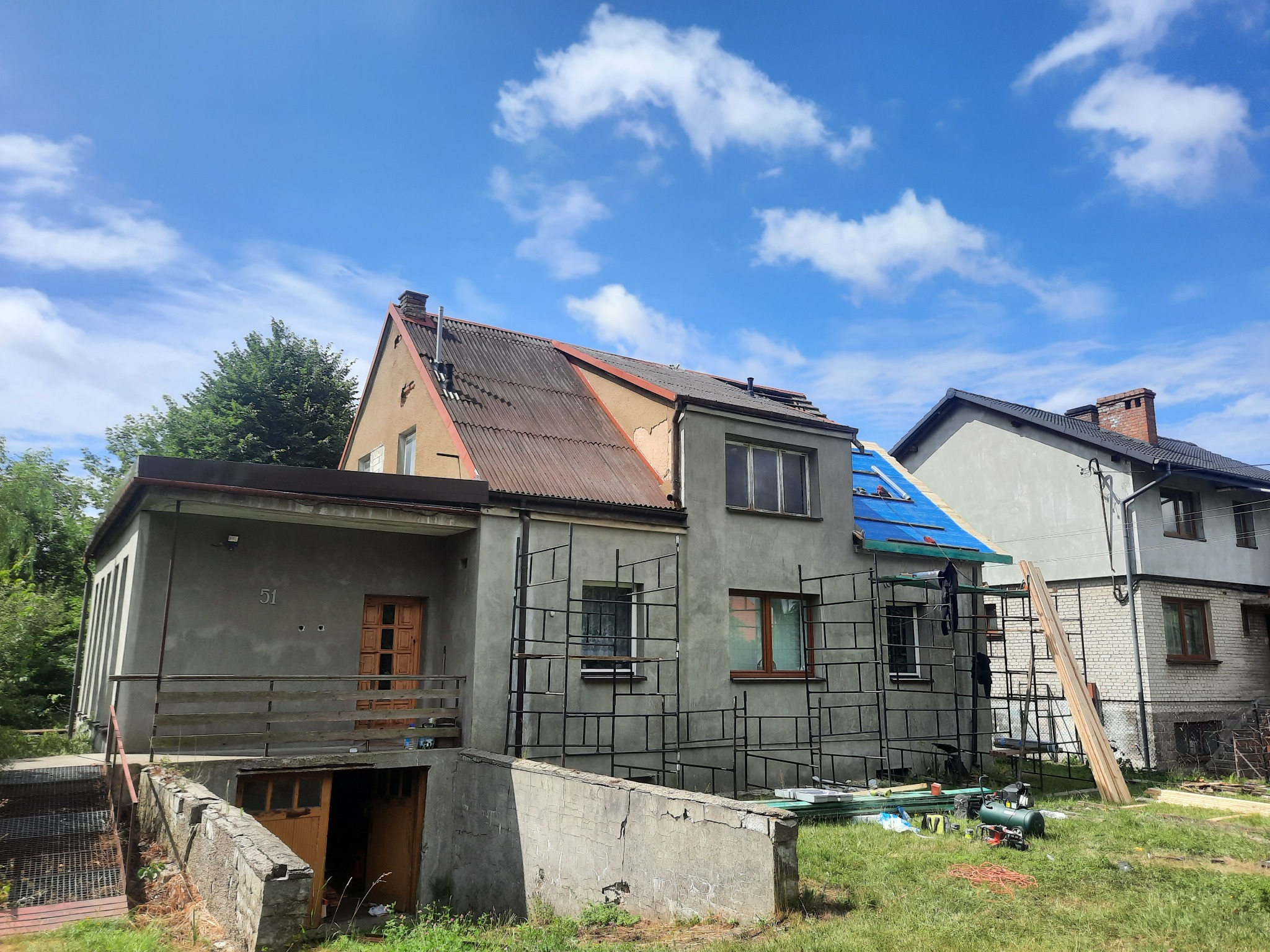 Dom w trakcie prac dekarskich, widoczne rusztowania, częściowo zdjęte stare pokrycie, niebieska plandeka zabezpieczająca, materiały budowlane na trawniku.