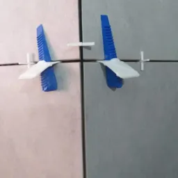 Szare płytki ścienne z niebiesko-białymi klinami poziomującymi i białymi krzyżykami dystansowymi, ujęcie z bliska.