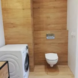 Nowoczesna łazienka z drewnopodobnymi płytkami na ścianach i podłodze, wisząca toaleta z chromowanym przyciskiem spłukiwania, biała pralka i fragment szafki z drewnianym blatem.
