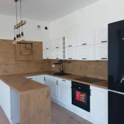 Nowoczesna kuchnia z białymi szafkami, drewnianym blatem i panelem ściennym, czarną armaturą i oświetleniem w stylu loft.