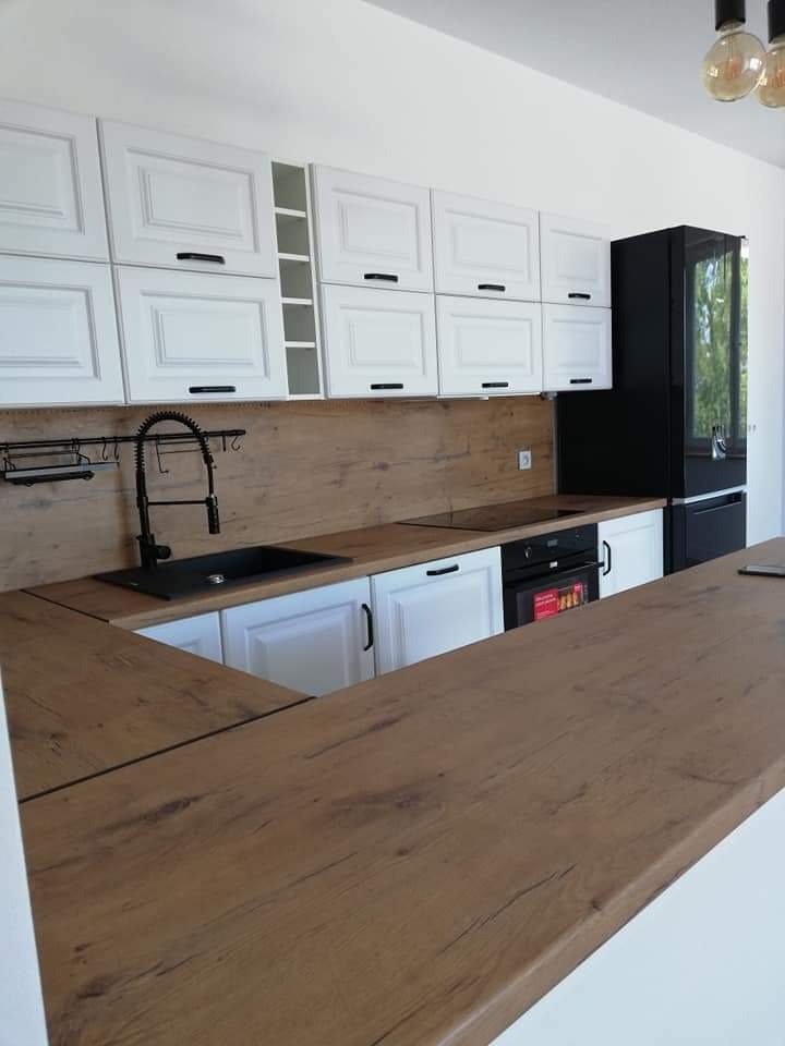Nowoczesna kuchnia z białymi szafkami z frezowanymi frontami, drewnianym blatem i czarnymi akcentami w postaci baterii i lodówki.