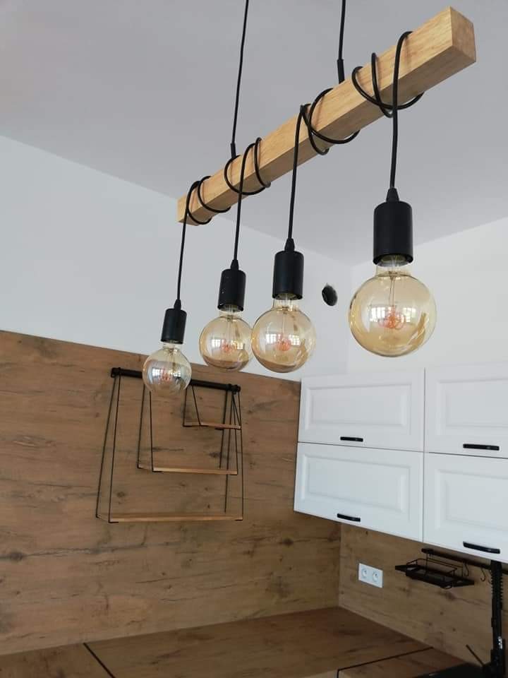 Lampa wisząca z drewnianą belką i czterema żarówkami Edison na tle białych szafek i drewnopodobnej okładziny ściennej z metalowymi półkami.