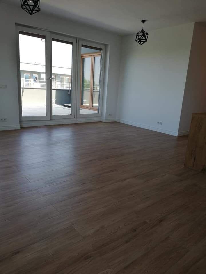 Jasne, puste pomieszczenie z parkietem w odcieniach brązu, duże okna balkonowe z widokiem na budynek naprzeciwko, nowoczesne, geometryczne lampy wiszące.