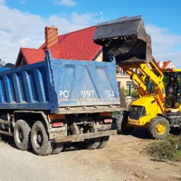 Żółta koparko-ładowarka JCB ładuje ziemię do niebieskiej ciężarówki na tle domów jednorodzinnych i błękitnego nieba.