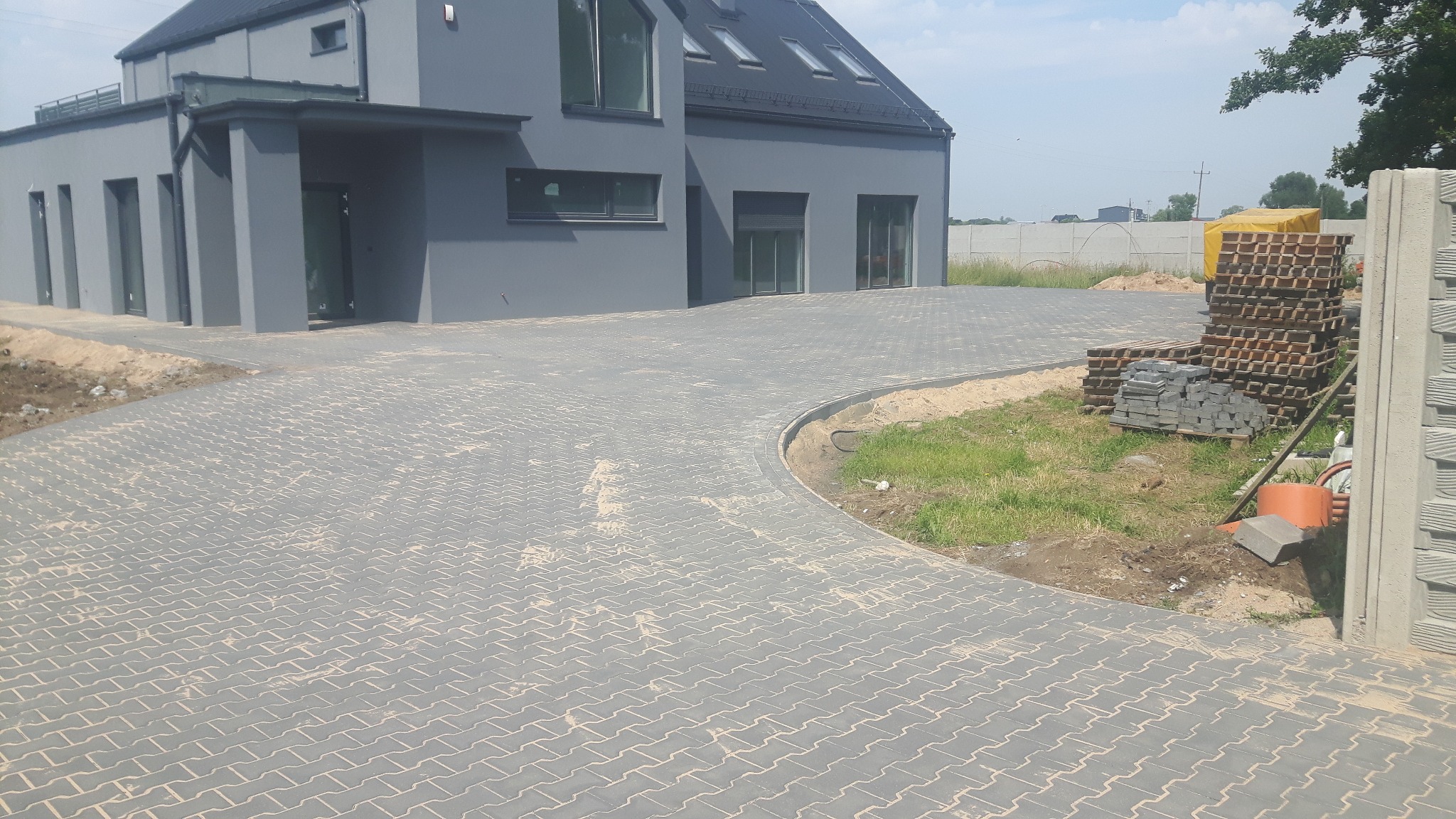 Nowoczesny dom z szarą elewacją i podjazdem wyłożonym szarą kostką brukową typu 'jodełka', widoczne palety z kostką i elementy obrzeży.