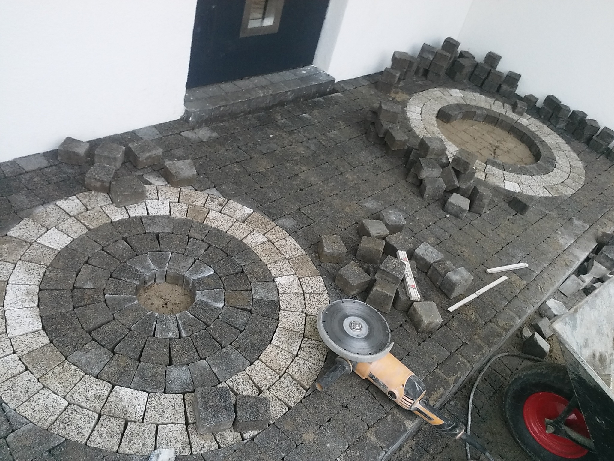 Fragment układanej nawierzchni z kostki brukowej w odcieniach szarości, z dekoracyjnymi okręgami z jasnej kostki, widoczna szlifierka kątowa i taczka z czerwonym kołem, etap prac.