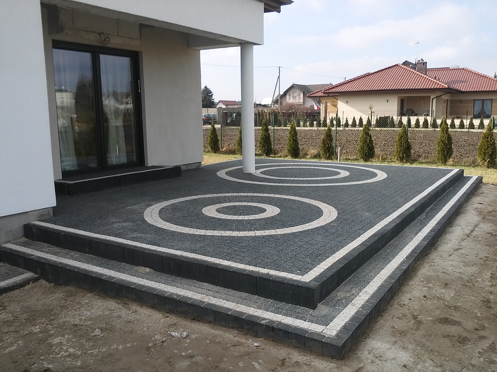 Nowoczesny taras z szarej kostki brukowej z białymi okręgami i schodami, widok z góry na dom i ogród.