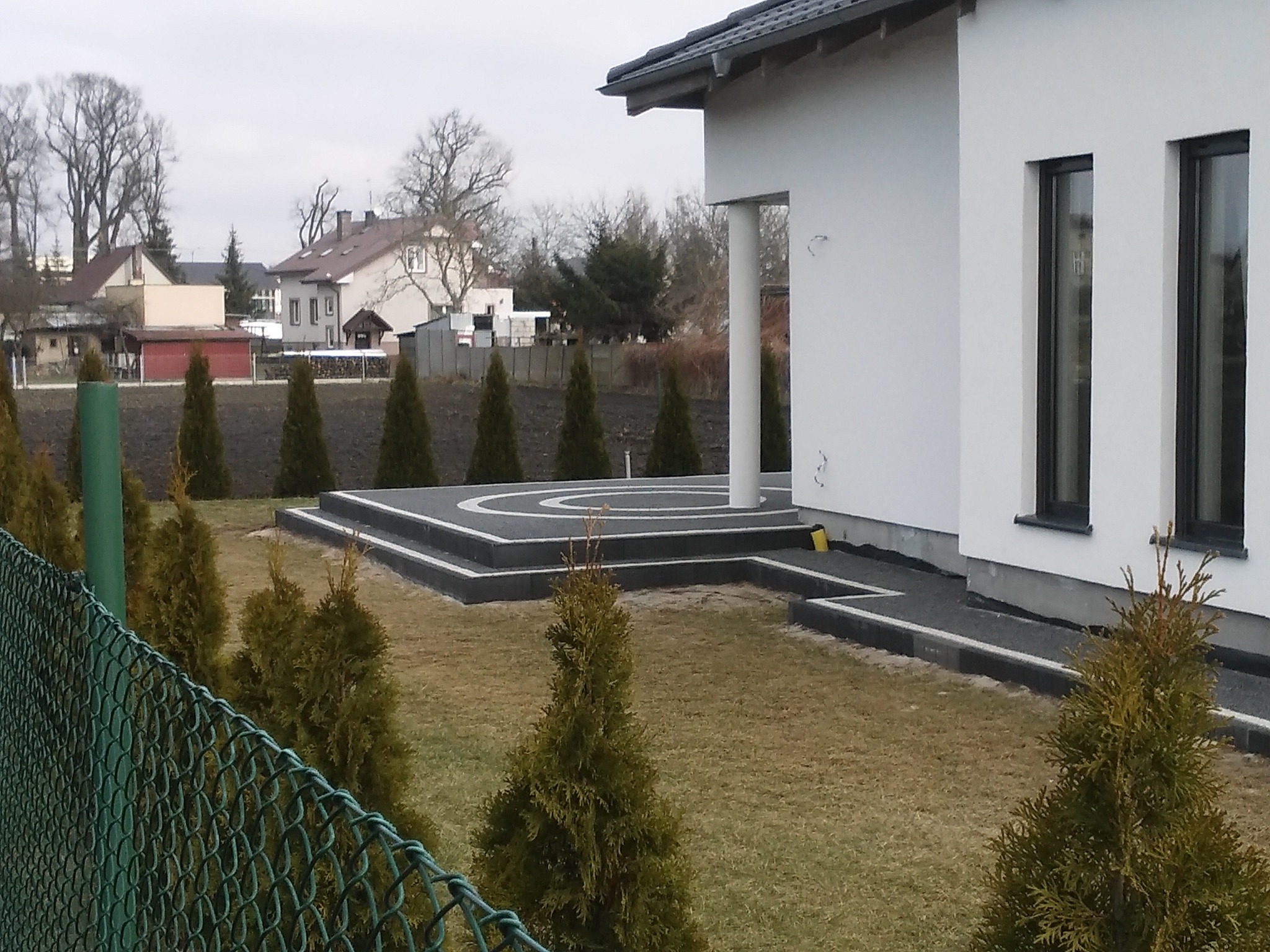 Ułożona kostka brukowa w kolorze czarnym z białym wzorem koła przed nowoczesnym domem, widok przez siatkę ogrodzeniową i małe tuje.