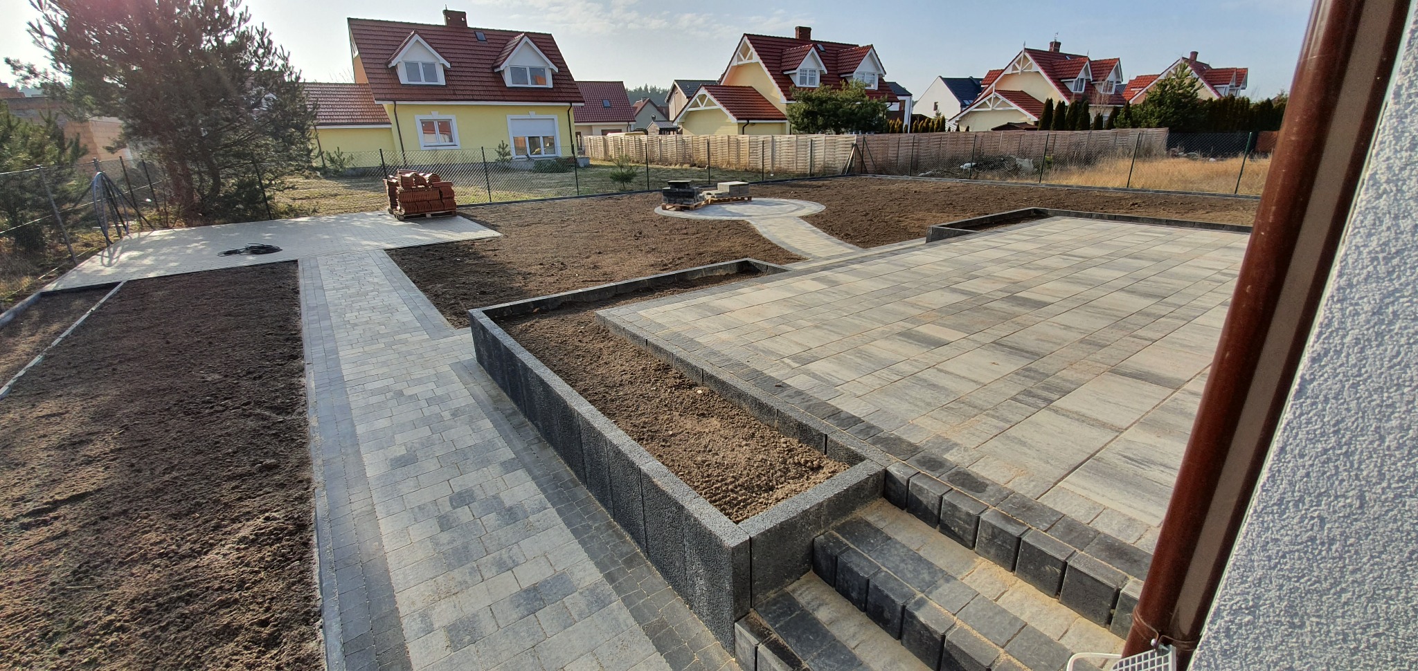Rozległy taras z kostki brukowej w odcieniach szarości i beżu, z niskimi obrzeżami z ciemnej kostki, otoczony przygotowanym pod zasiew trawnikiem i elementami ścieżek, w tle zabudowa jednorodzinna.