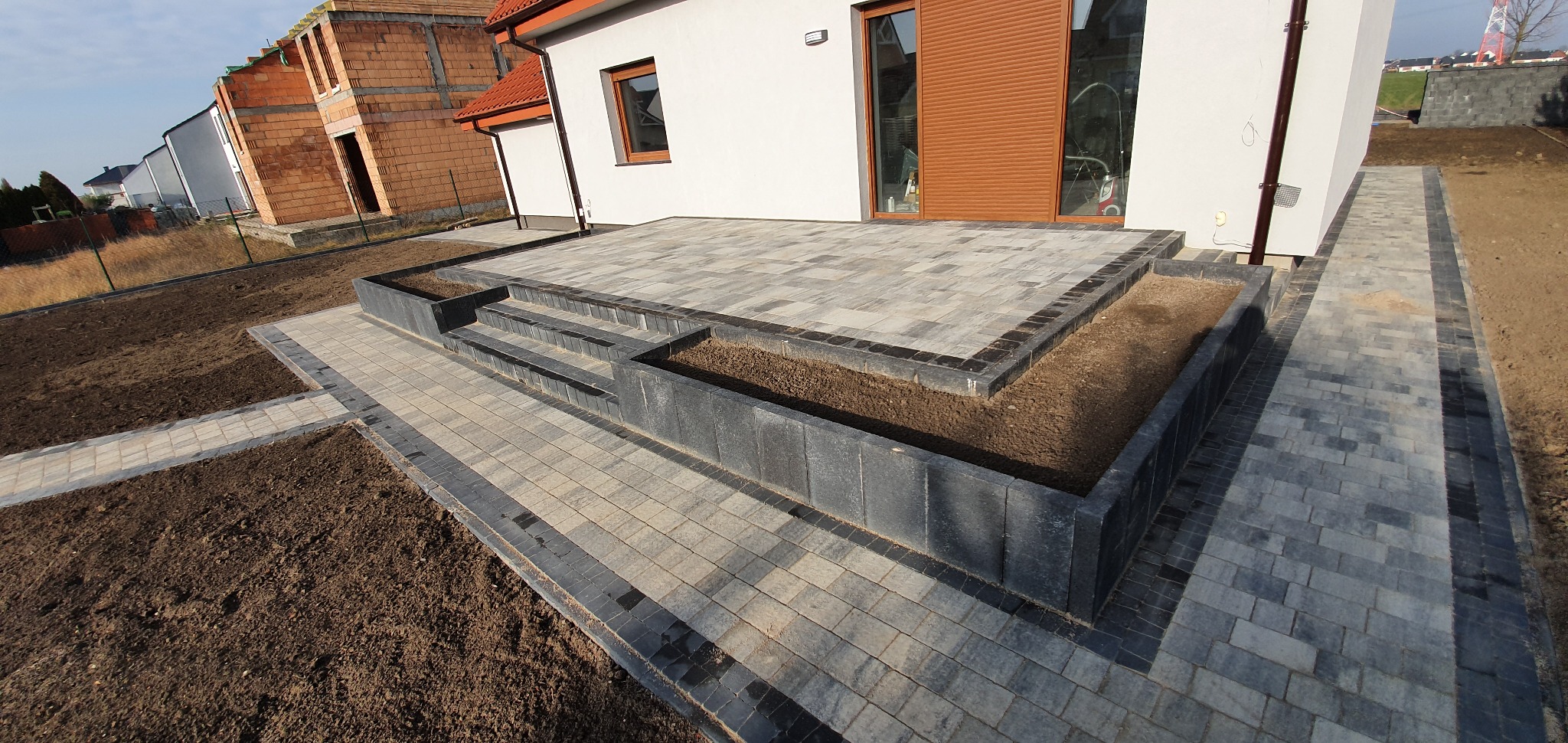 Nowoczesny taras z kostki brukowej w Poznaniu, z niskimi ściankami i schodami, otoczony przygotowanym terenem pod trawnik, widok na budynek w trakcie budowy.