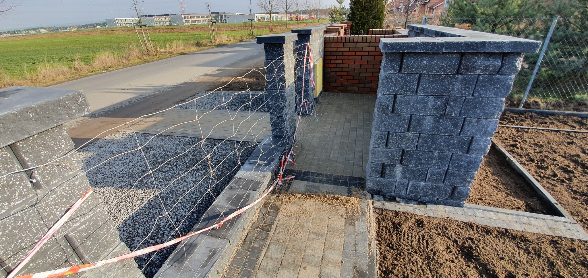 Fragment budowanego ogrodzenia z szarej kostki, widoczna siatka zabezpieczająca i częściowo ułożona nawierzchnia z kostki brukowej w odcieniach szarości i brązu, teren przygotowany pod dalsze prace.