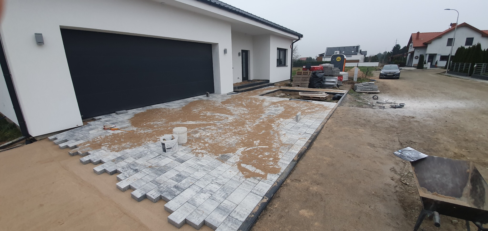 Niedokończony podjazd z szarej kostki brukowej ułożonej na piasku przed nowoczesnym domem z czarną bramą garażową. Część kostki już ułożona, widoczne białe wiadra z zaprawą i narzędzia. W tle inne...