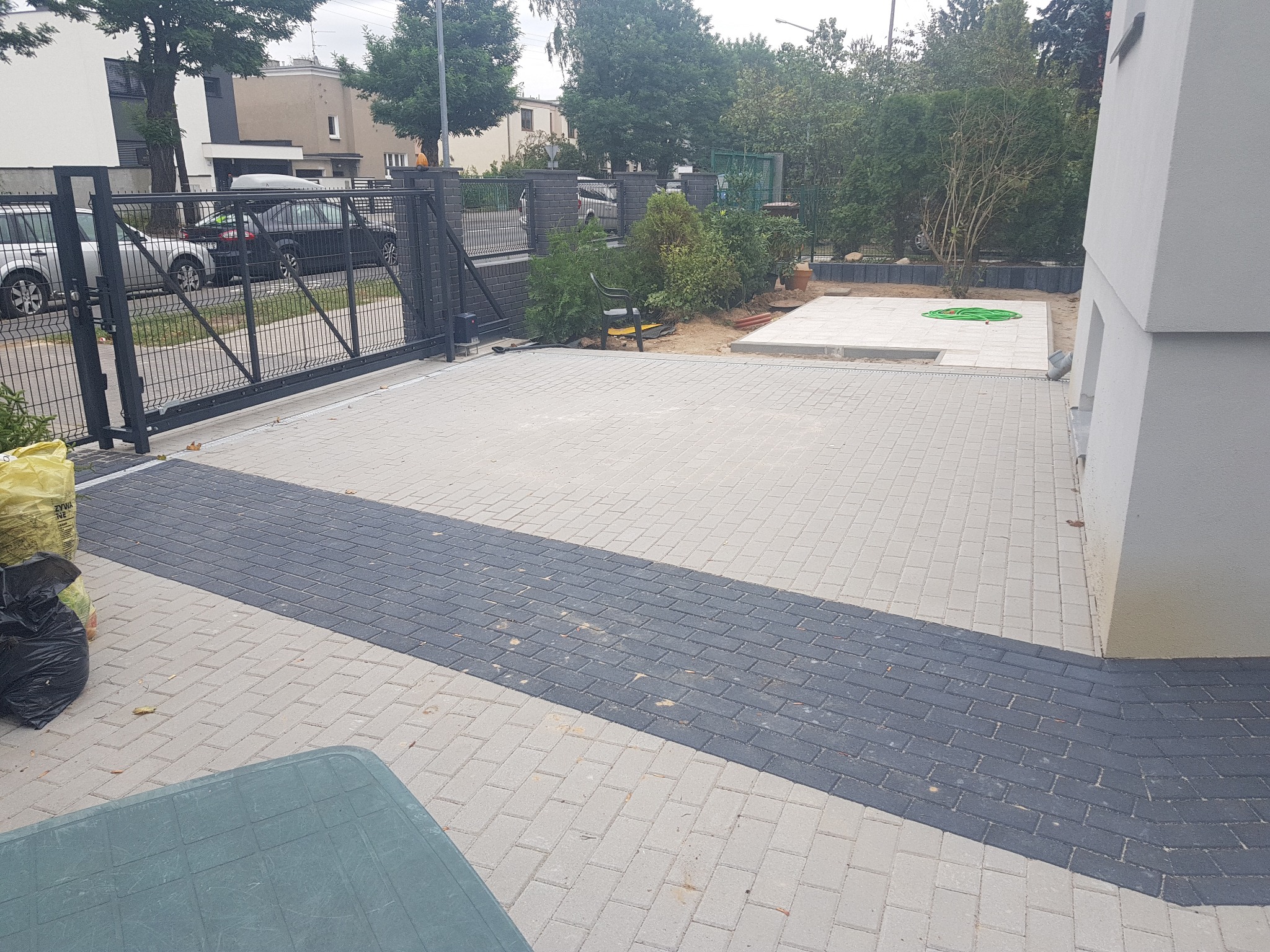 Ułożona jasnoszara kostka brukowa z ciemnoszarym pasem wzdłuż podjazdu, brama wjazdowa w tle, fragment budynku po prawej stronie, wąż ogrodowy zwinięty na betonowej platformie w głębi.