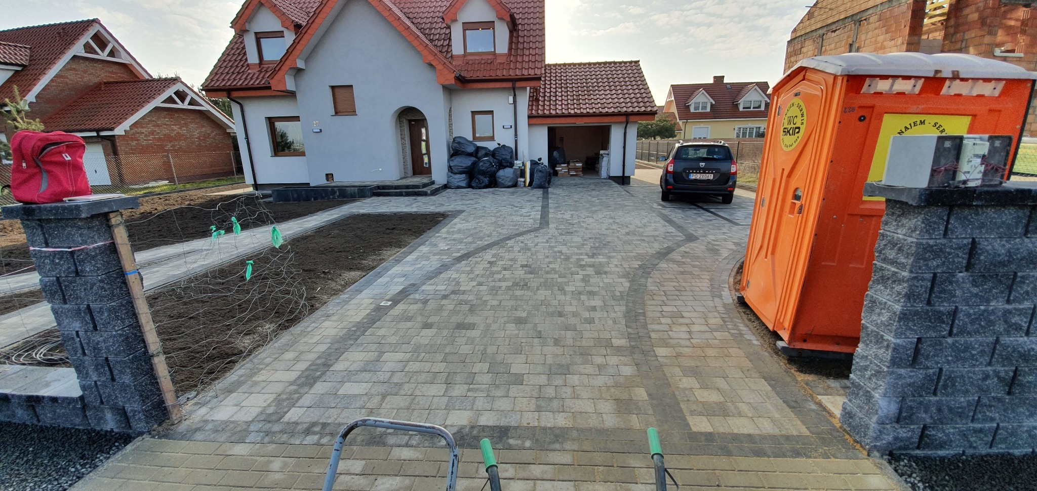 Nowo ułożona, wzorzysta nawierzchnia z kostki brukowej prowadząca do domu, widoczny fragment trawnika z siatką zabezpieczającą, przenośna toaleta i zaparkowany samochód.