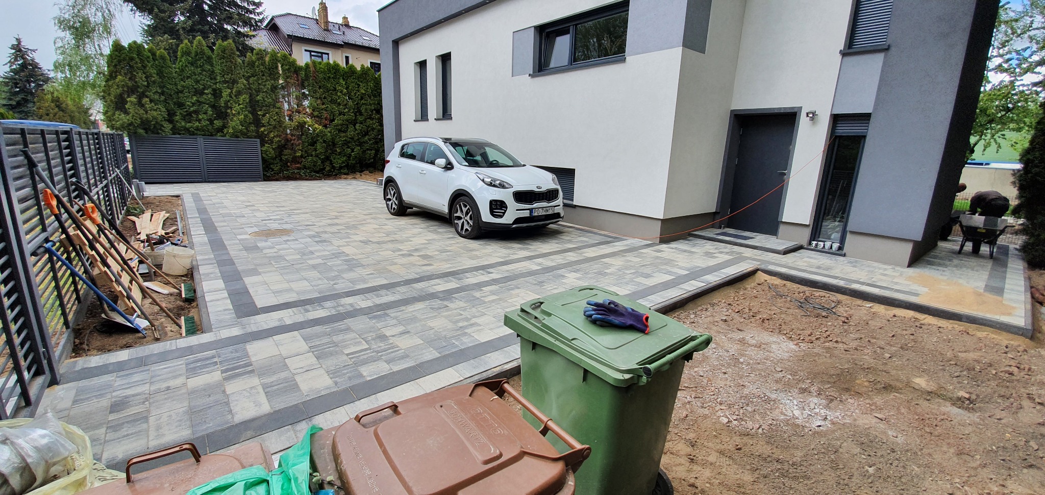 Ułożona kostka brukowa na podjeździe z samochodem Kia Sportage, widoczne kosze na śmieci i narzędzia brukarskie, częściowo wykończony teren.