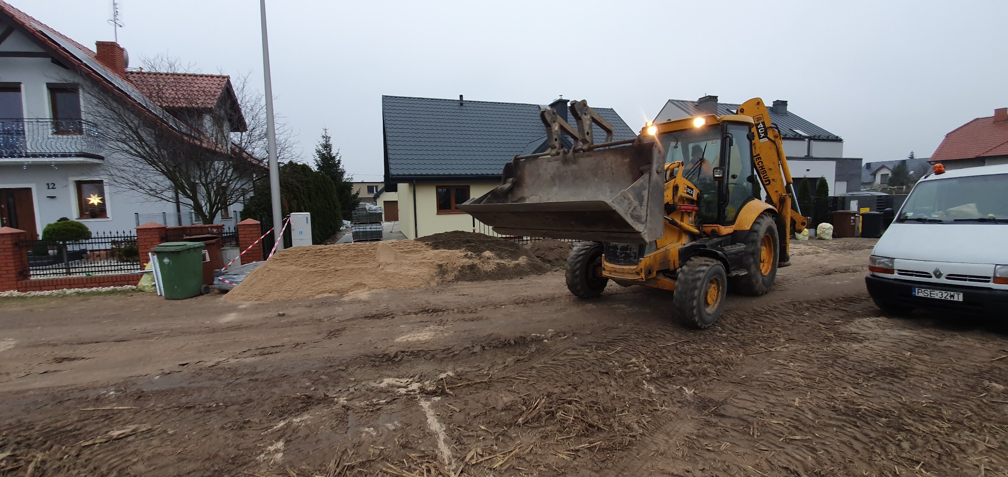 Żółta koparko-ładowarka JCB na placu budowy, z hałdą piasku i ziemi w tle, obok zaparkowany biały bus.