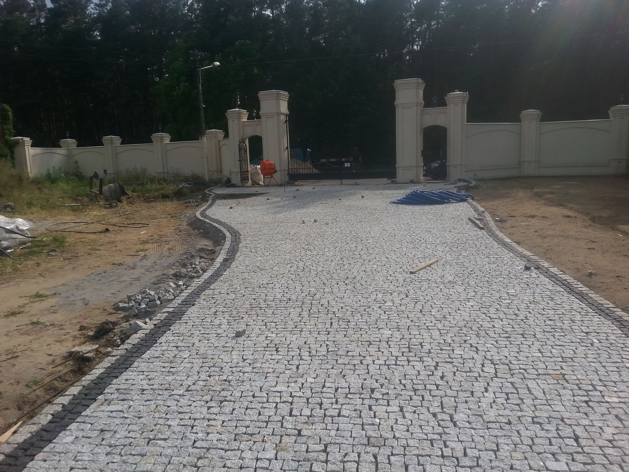 Szeroki, wybrukowany kostką granitową podjazd do posesji z ozdobnym ogrodzeniem i bramą w tle, widoczna betoniarka i materiały budowlane.