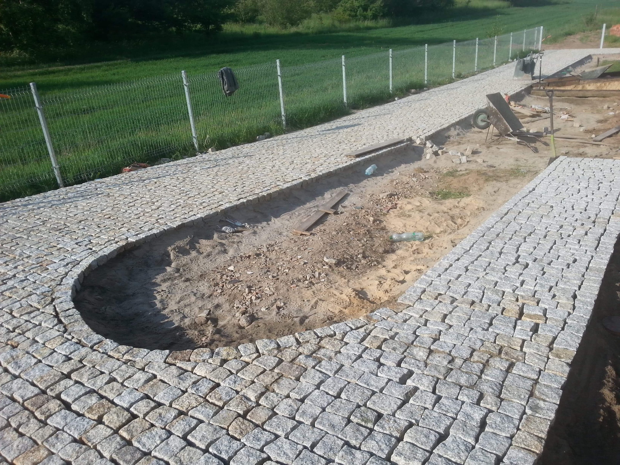 Fragment ścieżki z kostki granitowej w trakcie budowy, z widocznym wykopem i przygotowanym podłożem, w tle zielone pole i ogrodzenie z siatki.