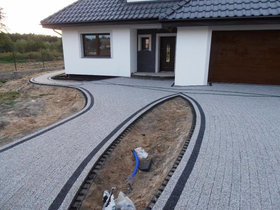 Szara kostka brukowa z czarnymi obramowaniami tworząca ścieżkę i podjazd przed nowoczesnym domem z brązową bramą garażową. Na środku widoczny fragment rabaty ziemnej z niebieskim przewodem.