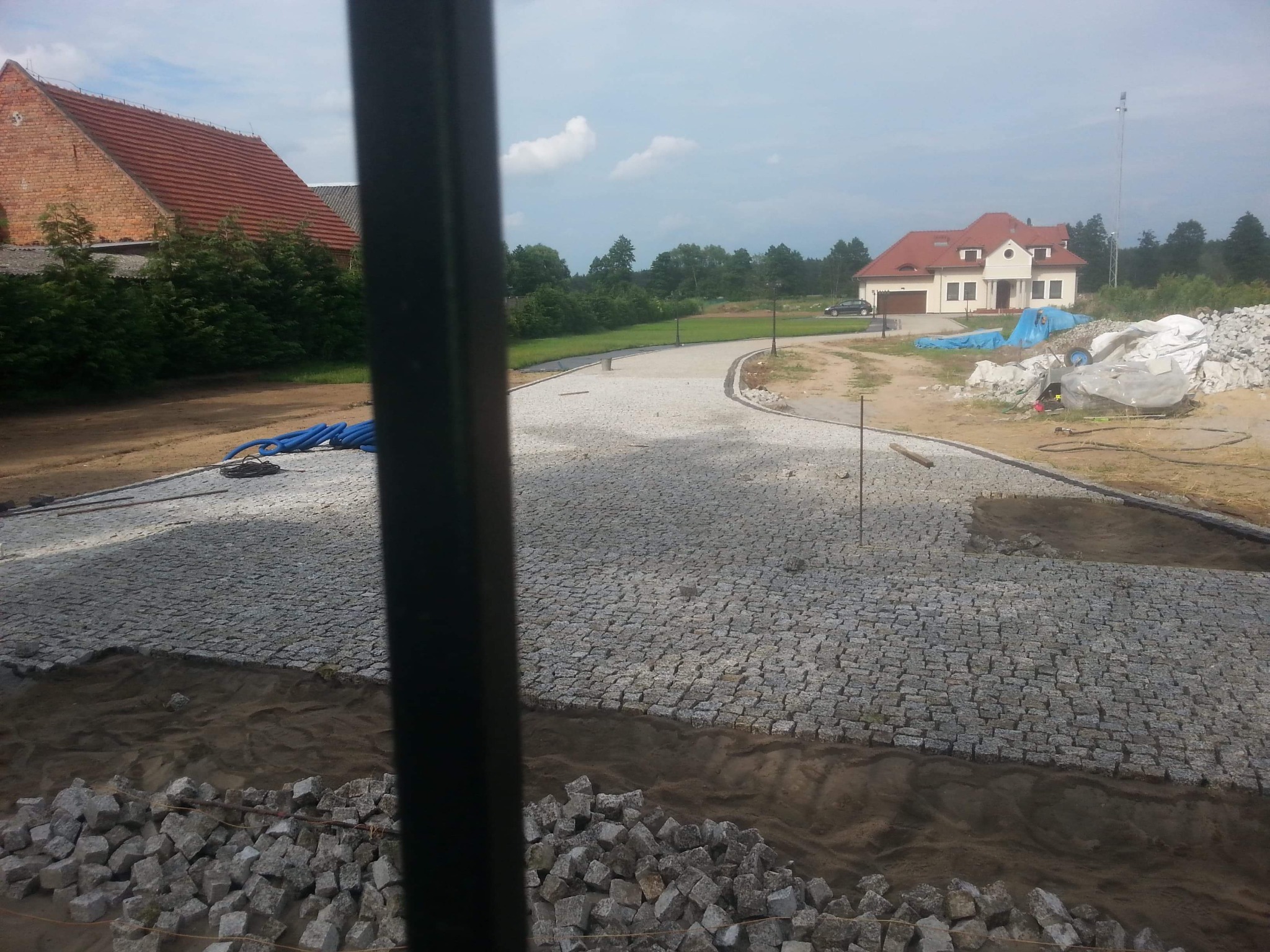 Fragment układanej drogi z kostki granitowej na posesji z dwoma domami, widok przez element konstrukcyjny.