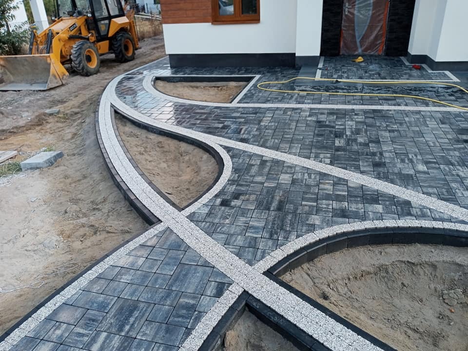 Układanie szarej kostki brukowej z białymi akcentami na podjeździe przed domem; w tle widoczna koparko-ładowarka i fragment elewacji budynku.