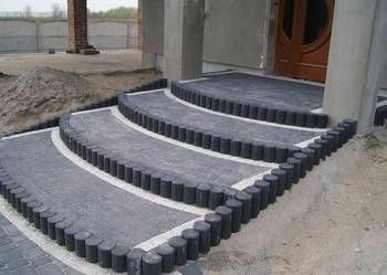 Schody wejściowe wyłożone szarą kostką brukową z obrzeżem z ciemnych, okrągłych palisad. W tle widoczny fragment domu w trakcie budowy i hałda piasku.