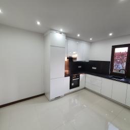 Marcin Łazarczyk - Nowoczesna, minimalistyczna kuchnia z białymi, lakierowanymi szafkami, czarnym blatem i widokiem na drzewo za oknem, oświetlona punktowymi lampami sufitowymi i z grzejnikiem żeberkowym w rogu.