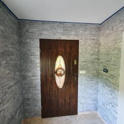 Marcin Łazarczyk - Przedpokój z dekoracyjną ścianą wykończoną szarą imitacją kamienia, drewniane drzwi wejściowe z owalnym przeszkleniem, widoczny panel sterowania na ścianie.