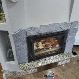 Otynkowany kominek z imitacją kamienia pomalowany na szaro, z farbą i pędzlem na podłodze, widoczny detal dekoracyjny w kształcie róży