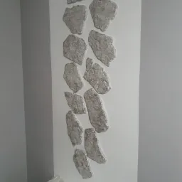 Dekoracyjna ściana z nałożoną strukturą przypominającą srebrne kamienie, ułożonych pionowo na białym tle, widoczny fragment narożnika pokoju pomalowanego na szaro.