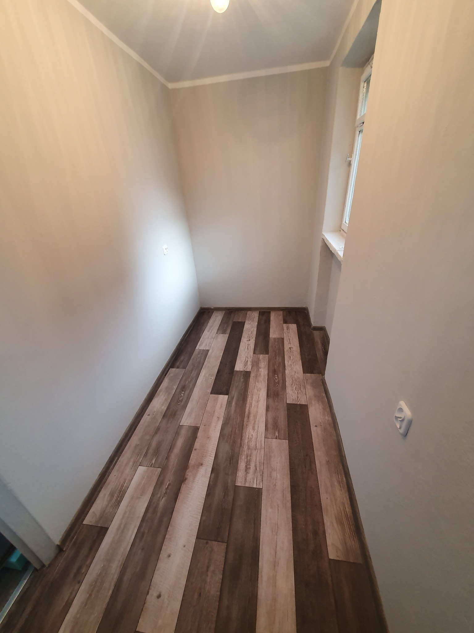 Wnętrze pokoju po remoncie z panelami podłogowymi imitującymi drewno ułożonymi wzdłuż, świeżo pomalowanymi ścianami w odcieniach szarości i nowym gniazdkiem elektrycznym na ścianie, widok na okno...